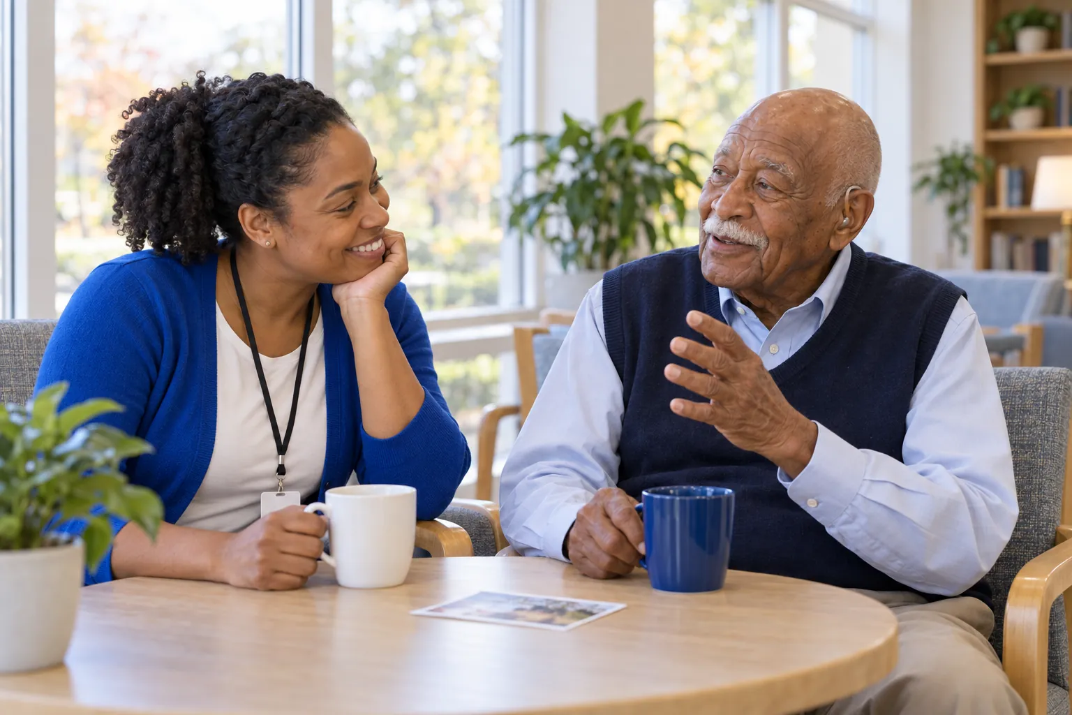 An activity assistant listens with a smile as a resident tells a story over coffee at a small table by the window